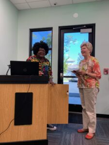 One person stands at a podium behind a computer, smiling. To their left is another person, smiling and holding a notebook.