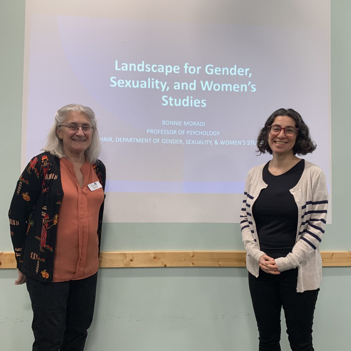 Two women stand in front of a projected presentation slide, reading, "Landscape for Gender, Sexuality, and Women's Studies".