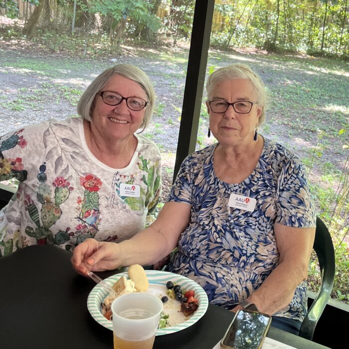 Two women sitting in a screened in porch with a plate of food. Woman on the left has short gray hair, woman on right has gray hair put up.