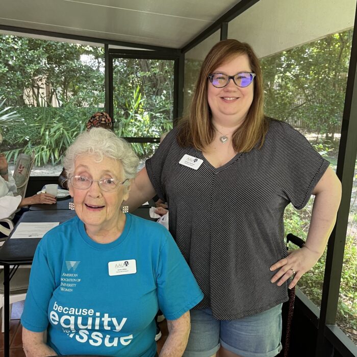 Two women in a screened in porch. Woman on the left with curly white hair seated, woman on the right with straight brown hair standing. Both smiling.